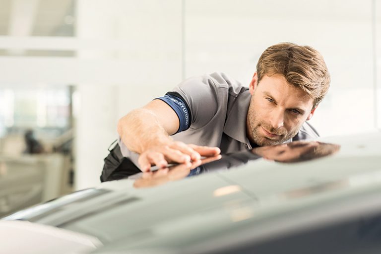 Servicetechniker beugt sich in einer Werkstatt über die Motorhaube eines Fahrzeugs und poliert sie mit einem weißen Tuch.