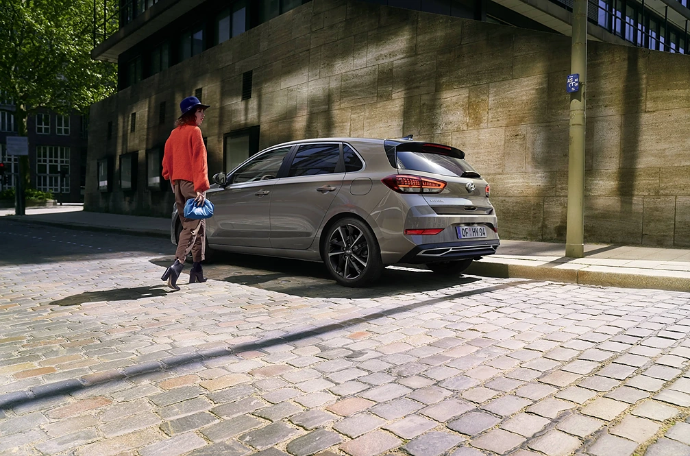 Frau mit rotem Pullover und Hut geht bei Tageslicht auf einen am Straßenrand geparkten silbernen Hyundai i30 zu; im Hintergrund ein modernes Bürogebäude.