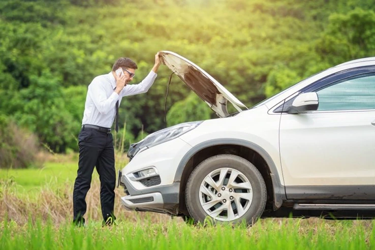 Mann steht auf einer Wiese neben einem silbernen SUV mit offener Motorhaube und telefoniert, vermutlich wegen einer Autopanne.