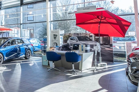 Verkaufsbereich in einem modernen Autohaus mit Schreibtisch, drei blauen Stühlen und rotem Sonnenschirm; umgeben von ausgestellten Fahrzeugen vor großer Fensterfront.