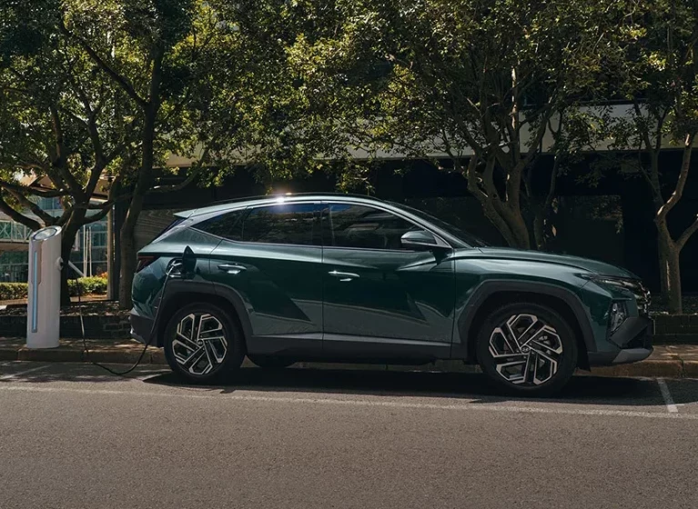Grüner Hyundai TUCSON Plug-in-Hybrid steht auf einem Parkplatz unter Bäumen und ist an eine Ladesäule angeschlossen.