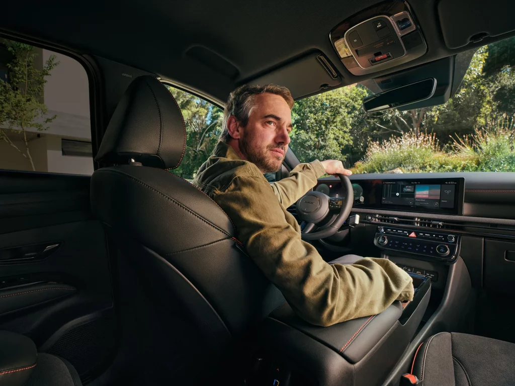 Innenansicht des Hyundai TUCSON mit digitalem Cockpit, Touchscreen und einem Mann am Steuer bei Tageslicht.