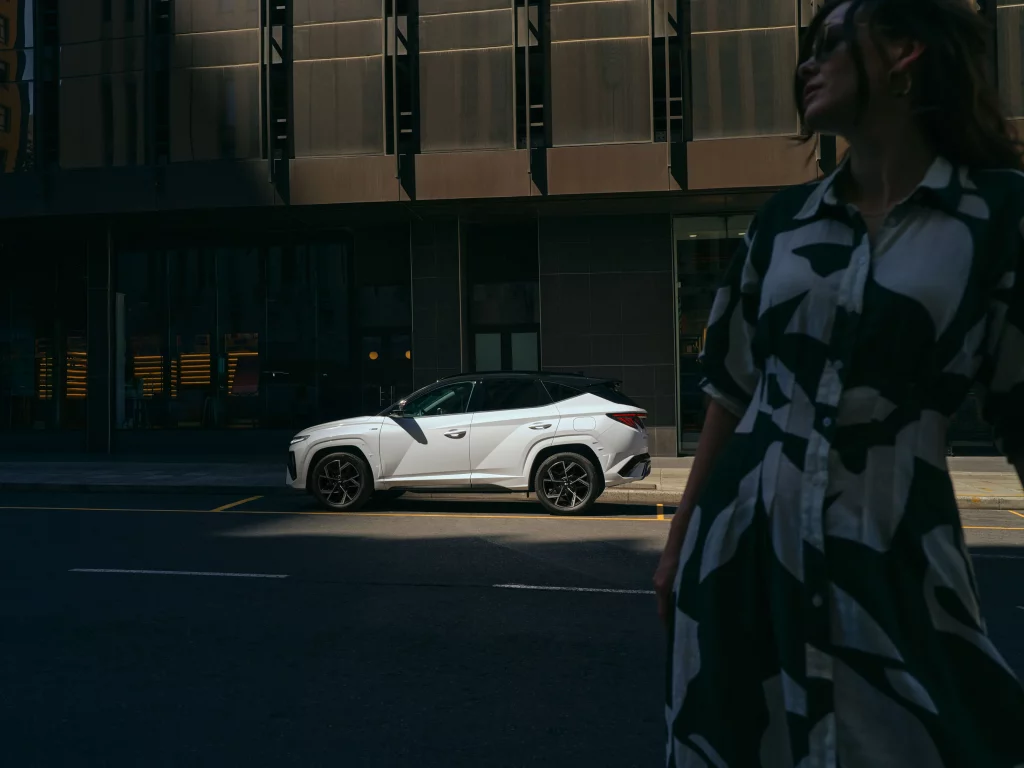 White Hyundai TUCSON parked at the curb in front of a modern high-rise building; a stylishly dressed woman stands in the shadowed foreground