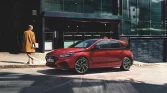 Roter Hyundai Kompaktwagen seitlich vor modernem Glasgebäude, Frau im gelben Mantel auf Bürgersteig