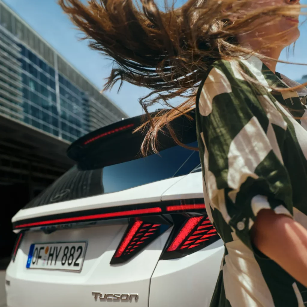 Weißer Hyundai SUV von hinten vor modernem Glasgebäude, Frau mit wehenden Haaren in urbaner Umgebung