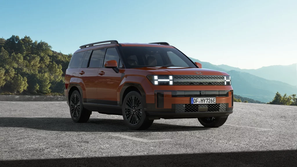 Orangener moderner SUV seitlich auf Asphalt mit grüner Landschaft und Bergen im Hintergrund geparkt