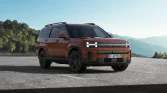 Orangener moderner SUV seitlich auf Asphalt mit grüner Landschaft und Bergen im Hintergrund geparkt