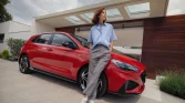 Frau lehnt an rotem Hyundai Kompaktwagen vor modernem Haus mit Holzfassade und großer Einfahrt
