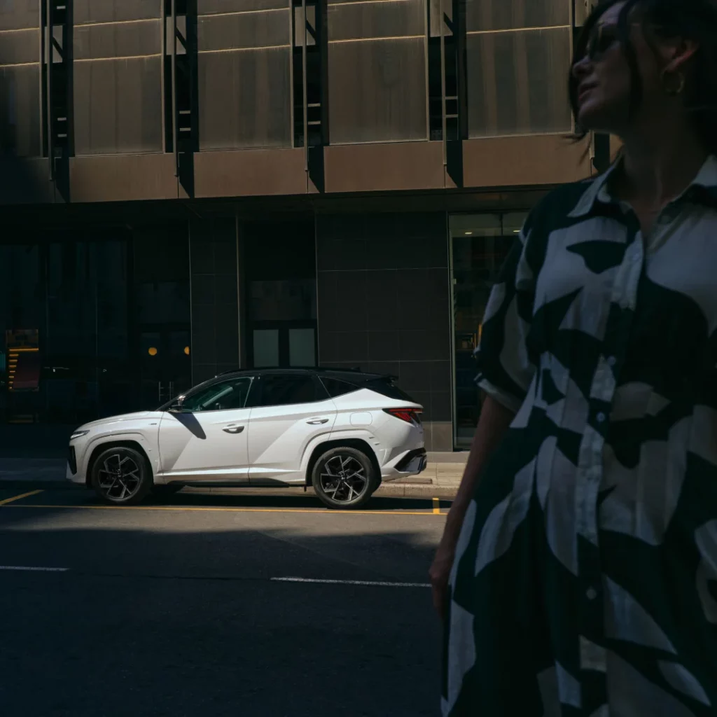 Weißer moderner SUV geparkt vor dunklem Bürogebäude, Person mit gemustertem Outfit im Schatten sichtbar