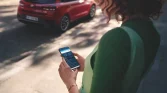 Frau mit Smartphone neben rotem Hyundai Kompaktwagen, städtischer Sonnenschein und Gehweg im Hintergrund