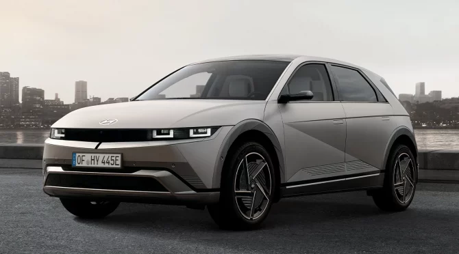 Silberner Hyundai Elektro-SUV seitlich am Wasser mit moderner Stadt im Hintergrund bei Sonnenuntergang