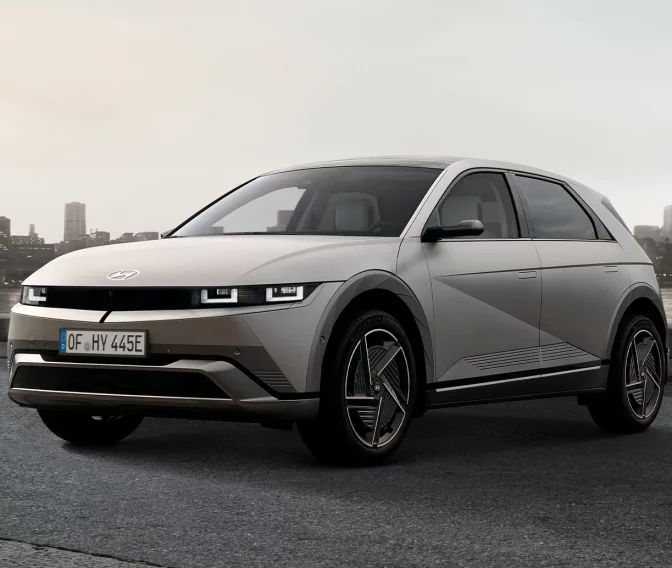 Silberner Hyundai Elektro-SUV seitlich am Wasser mit moderner Stadt im Hintergrund bei Sonnenuntergang
