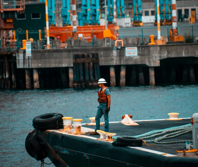 Mann mit Schutzhelm auf Schlepperdeck vor industriellem Hafen mit Kränen und Lagerhallen im Hintergrund