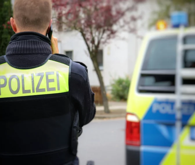 Polizeibeamter mit Warnweste von hinten, telefonierend vor grün-blauem Polizeivan in Wohnstraße im Herbst