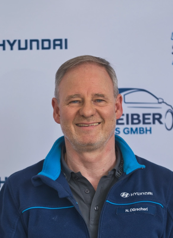 Männlicher Hyundai-Mitarbeiter in blauer Jacke vor Unternehmenslogo, freundlich lächelnd und professionell posierend