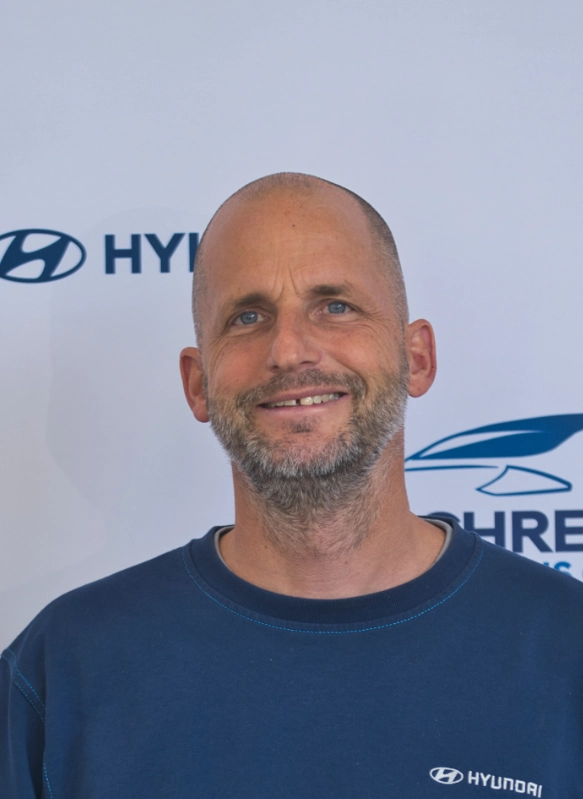 Männlicher Hyundai-Mitarbeiter mit Bart lächelt vor weißem Hintergrund mit Hyundai-Logo