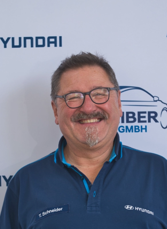 Mann mit Brille und blauem Hyundai-Hemd lächelnd vor Firmenlogo auf weißem Hintergrund