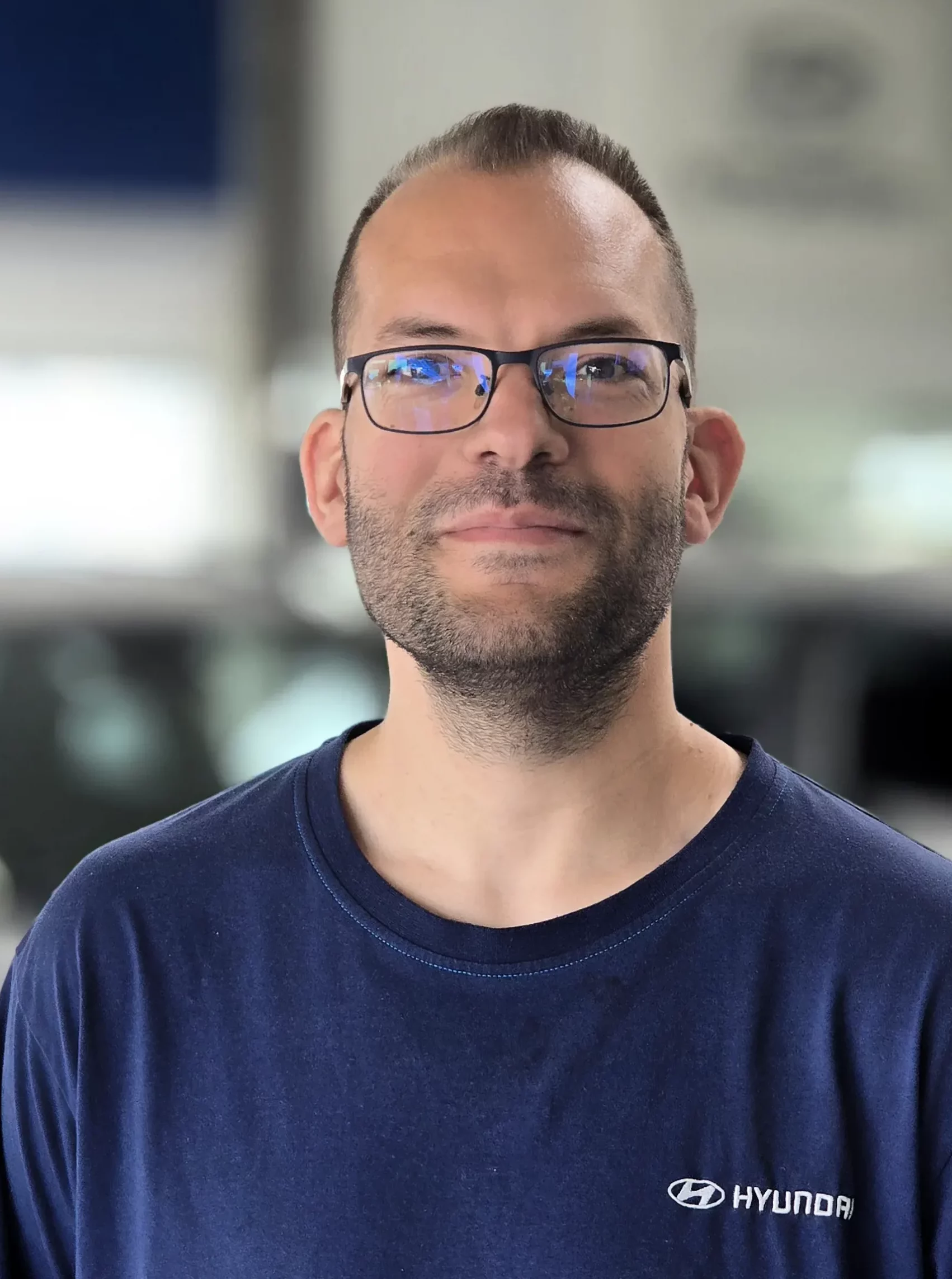 Porträt eines Mannes mit dunkelblauem Hyundai-Shirt, Brille und Bart in Autohaus-Umgebung — Vollinger